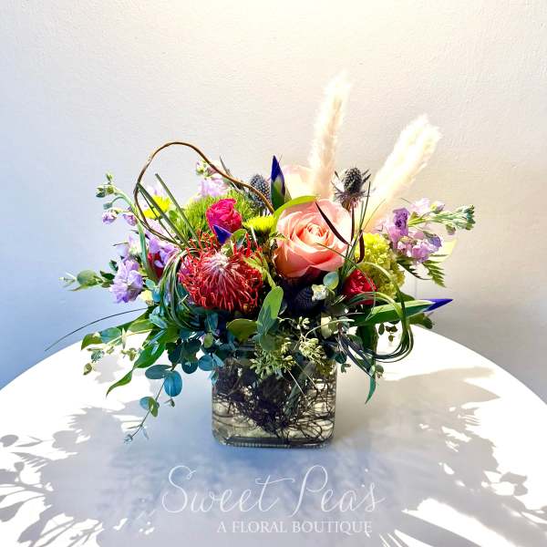 Mixed floral arrangement in a square glass vase with a pink rose and purple blooms
