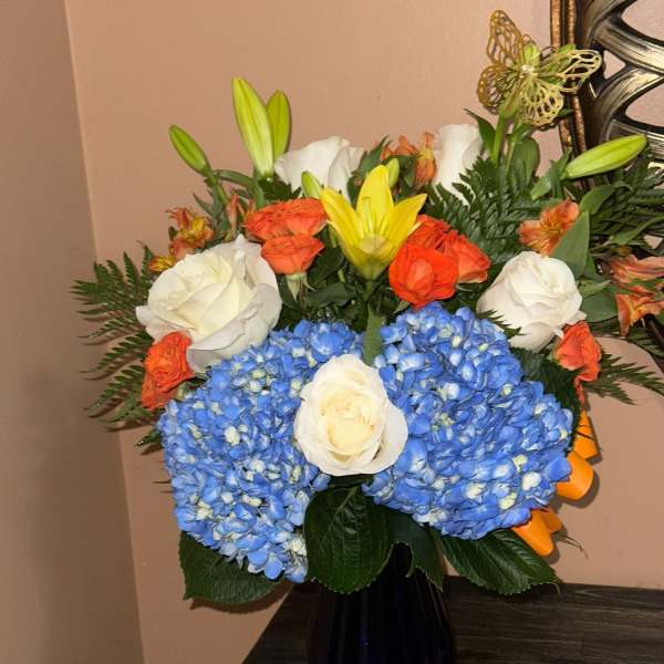 Bouquet of blue hydrangeas, white roses, orange roses, and yellow lilies in a dark vase