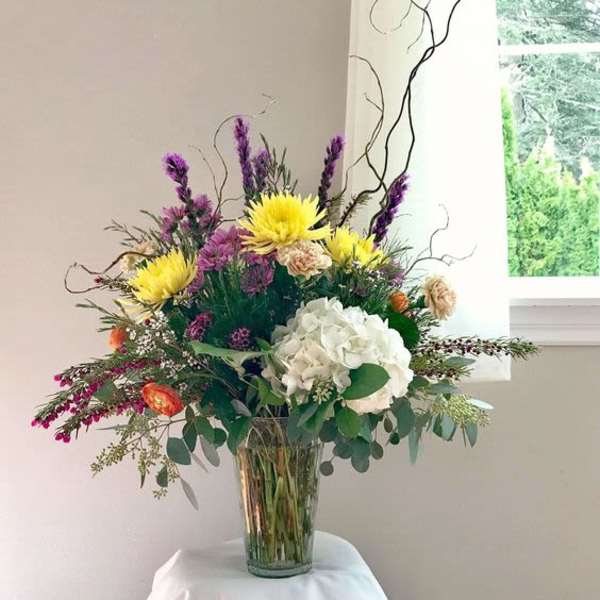 Tall mixed arrangement with yellow mums, white hydrangea and purple flowers in a clear glass vase