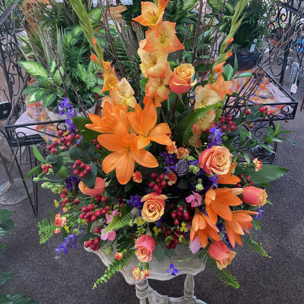 Tall arrangement of orange lilies and gladiolus with peach roses and purple flowers in a mixed display