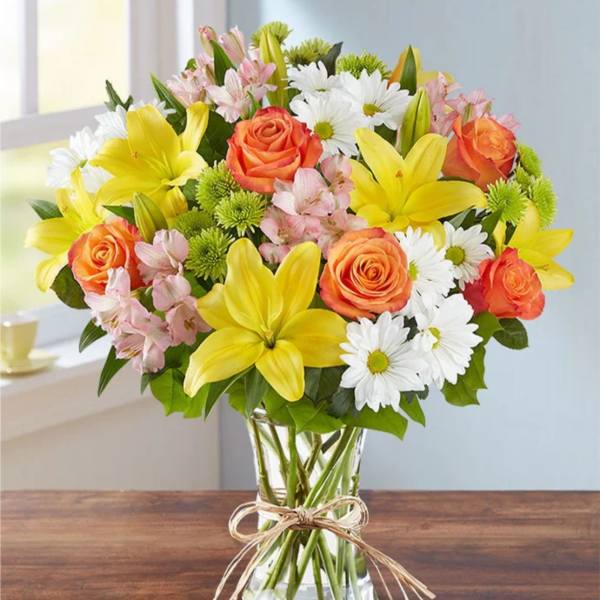 Bouquet of yellow lilies, orange roses, and white daisies in a glass vase