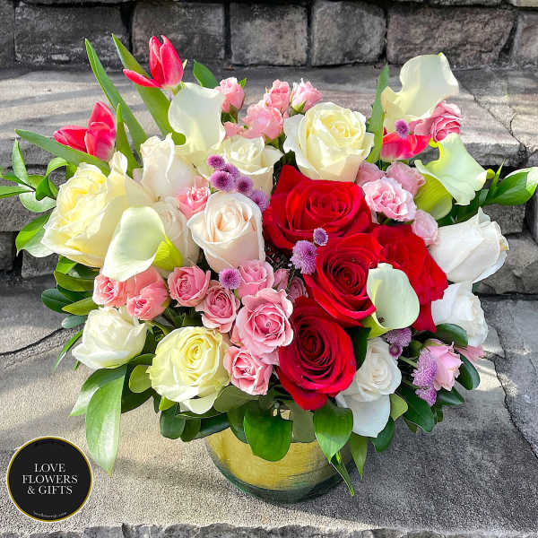 Round arrangement of red and white roses, pink spray roses, and white calla lilies in a glass vase.