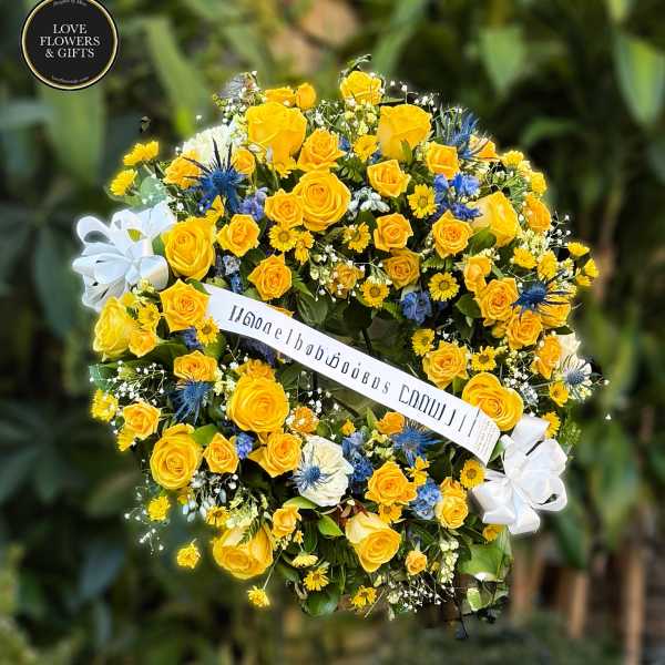 Round standing wreath of yellow roses and daisies with blue accents and a white ribbon banner.