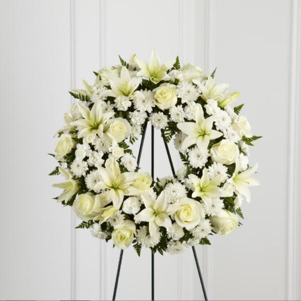 White floral wreath on a black stand with lilies and roses