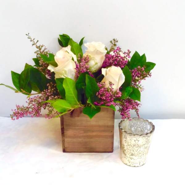 White roses and pink filler flowers in a wooden box with a glass votive beside it