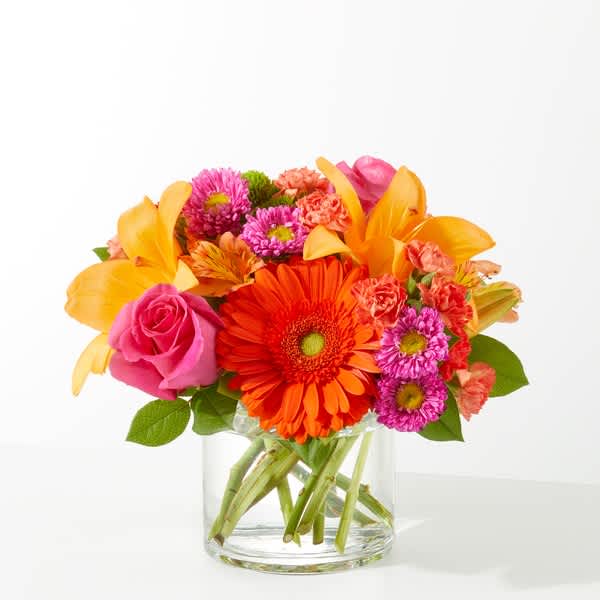 Bright mixed arrangement of orange gerbera, lilies, pink roses and small blooms in a clear glass cylinder vase