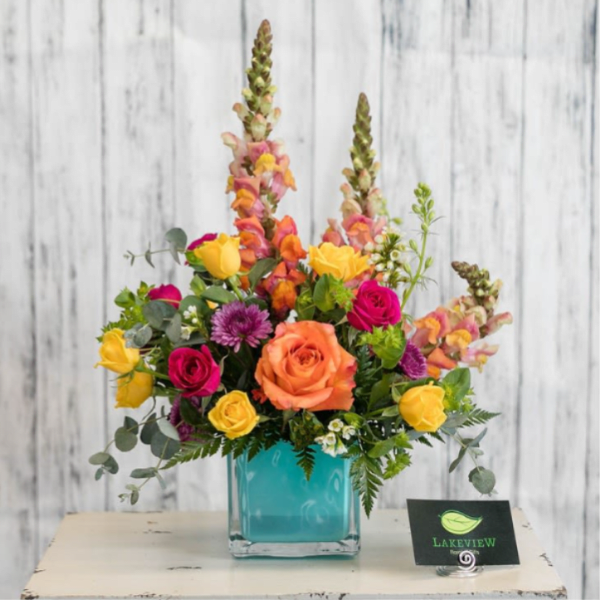Bright mixed arrangement with orange, yellow, and pink flowers in a turquoise cube vase