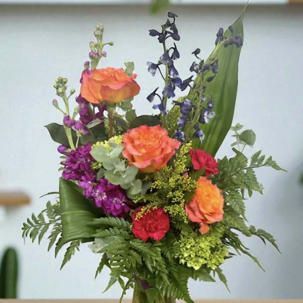 Mixed bouquet of orange roses, purple flowers, and greenery in a glass vase