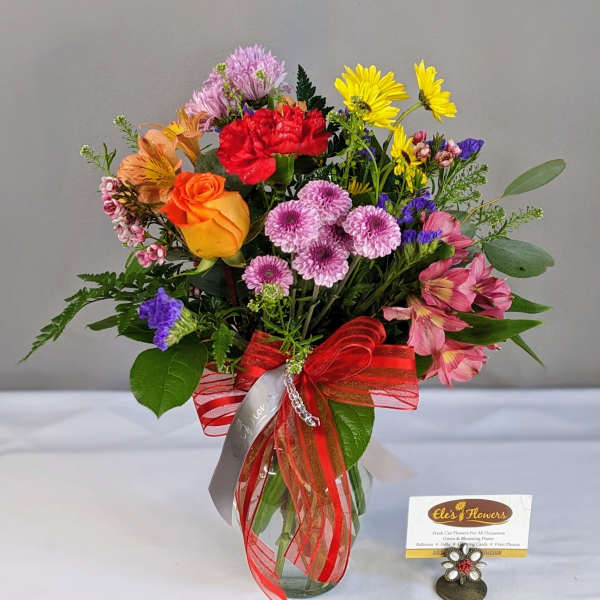 Bright mixed bouquet of roses, carnations, daisies, and mums in a clear vase with a red bow.