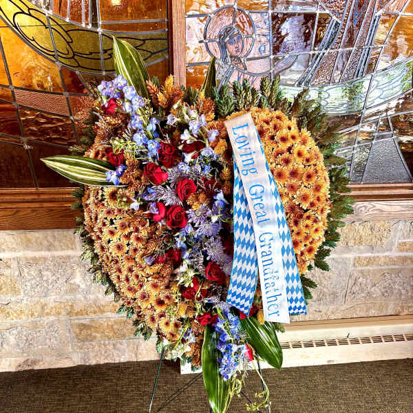 Heart-shaped standing funeral spray of orange mums with red roses, blue flowers, ribbon, and large green leaves on an easel
