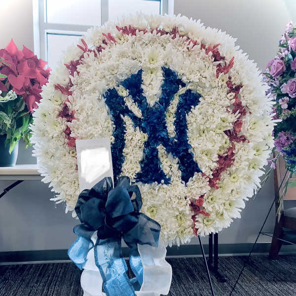Large round standing wreath of white flowers with red accents and a blue floral emblem, finished with a dark bow.