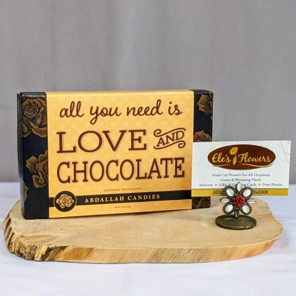 Box of assorted chocolates with love-themed text on a wooden board beside a floral card holder.