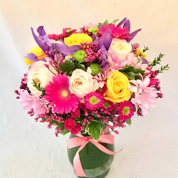 Bright mixed bouquet of pink, yellow, purple, and white flowers in a clear glass vase with a pink ribbon.