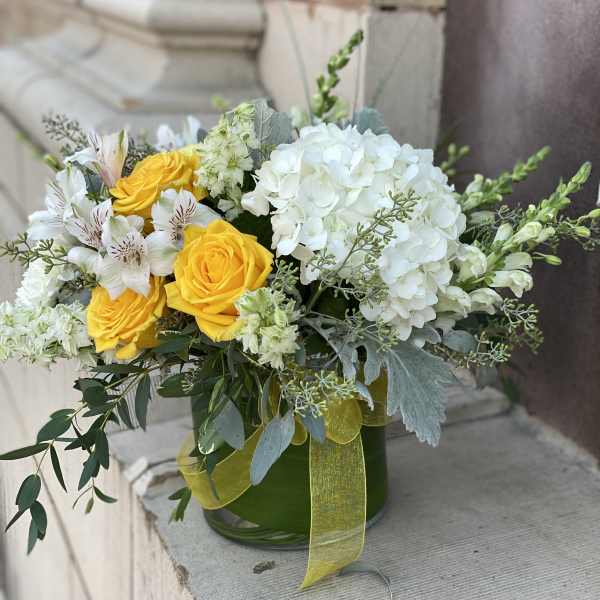Low glass vase of yellow roses, white hydrangeas, and white blooms with a yellow ribbon.