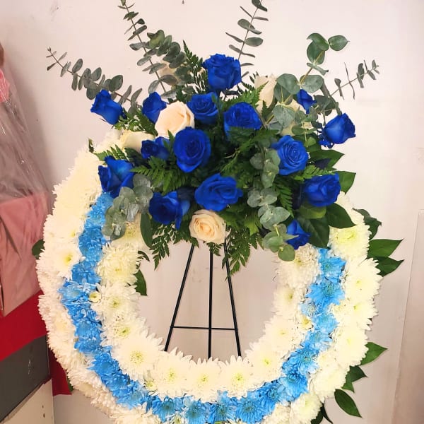 Blue and cream rose arrangement above a white-and-blue floral wreath on a stand