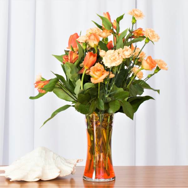 Tall arrangement of orange and peach roses, carnations, and other blooms in a clear glass vase with orange water.
