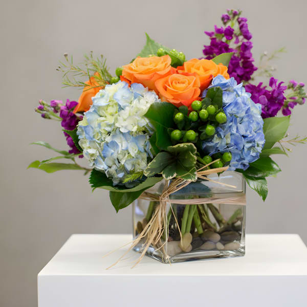 Bouquet of orange roses, blue hydrangeas, and purple flowers in a glass cube vase