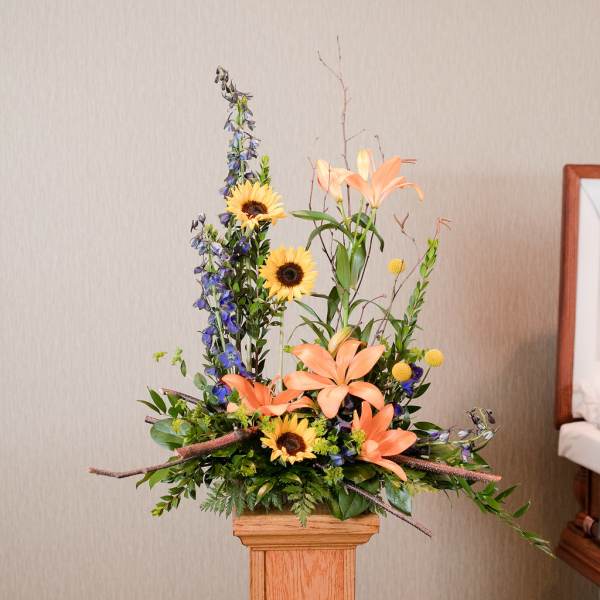 Tall floral arrangement with peach lilies, sunflowers, and blue delphinium in a wooden pedestal vase