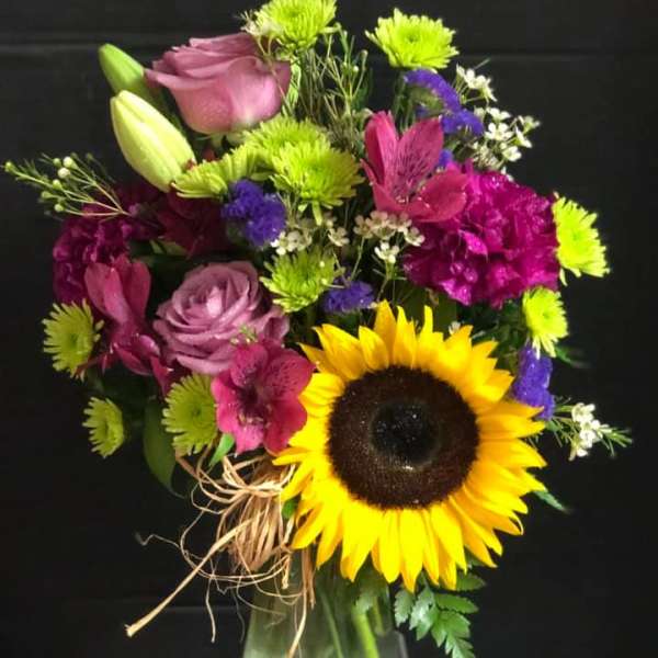 Mixed bouquet with a sunflower, roses, lilies, and purple blooms in a glass vase