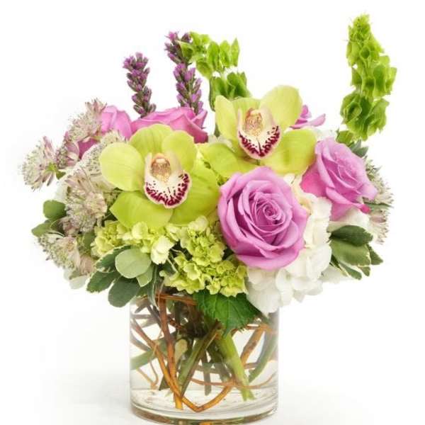 Short arrangement of green orchids, pink roses, and white blooms in a clear glass cylinder vase