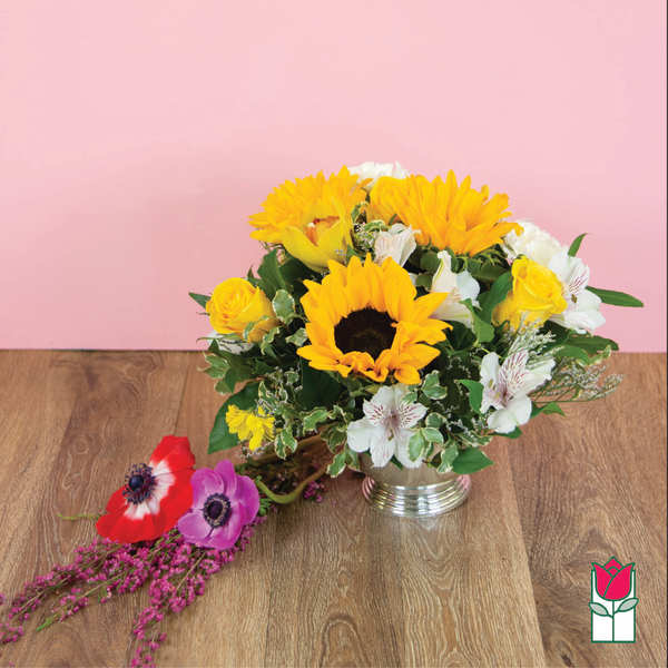 Yellow sunflowers and white flowers in a silver vase with loose blooms beside it.