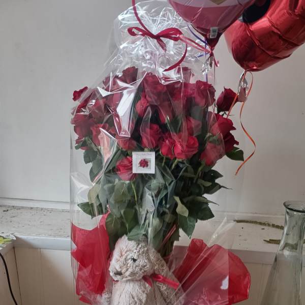 Bouquet of red roses wrapped in clear plastic with a teddy bear and heart balloons