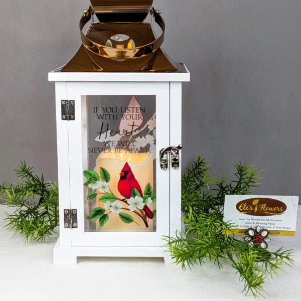 White lantern with a lit candle and cardinal design, surrounded by greenery.