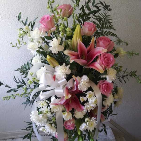 Standing floral spray with pink roses, lilies, and white flowers tied with a white ribbon