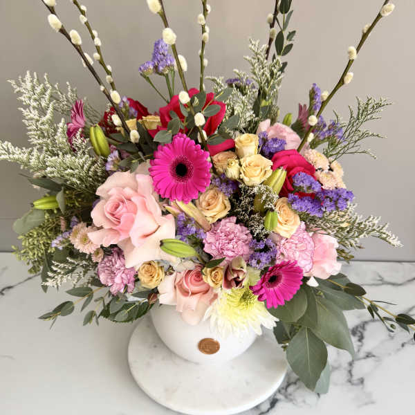 Colorful arrangement of pink gerberas, roses and mixed blooms in a white round vase with tall pussy willow branches