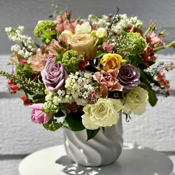 Mixed arrangement of cream, peach, lavender, and yellow roses with small blooms in a white ceramic vase