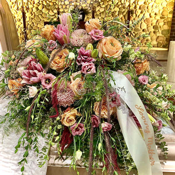 Large peach and pink casket spray with roses, lilies, and mums draping over a bronze casket