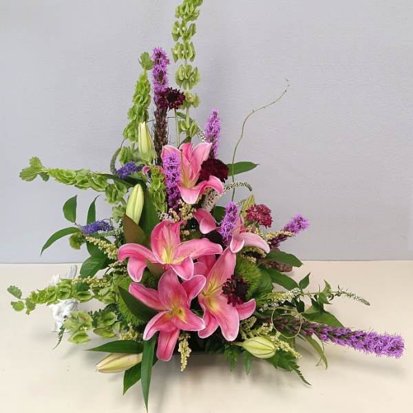Pink lilies arranged with tall green and purple flowers in a low container