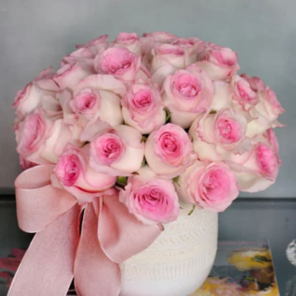Pink and white roses arranged in a white vase with a pink ribbon