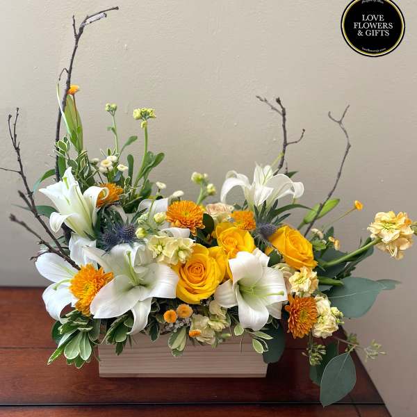 Low arrangement of yellow roses, white lilies, and orange mums in a rectangular wooden box.