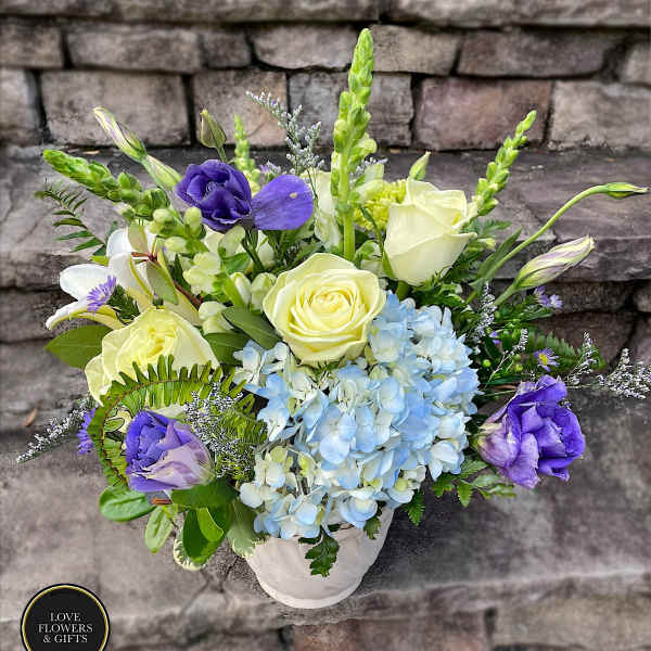 Arrangement of pale yellow roses, blue hydrangeas, and purple blooms in a white vase on stone steps