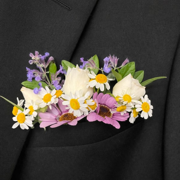 Floral boutonniere with white daisies, purple blooms, and cream rose buds on a black jacket