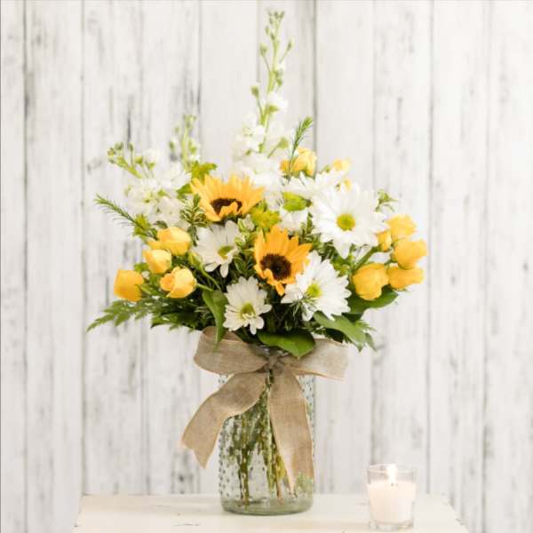 Yellow roses and white daisies in a glass vase with a burlap bow.
