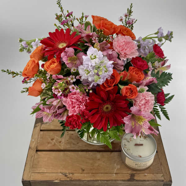 Bright mixed bouquet of red, pink, and orange flowers in a clear vase with a tin candle on a wooden crate