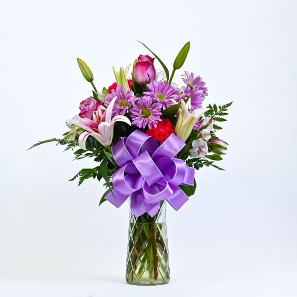 Bouquet of pink lilies, roses, and purple daisies in a glass vase with a purple bow