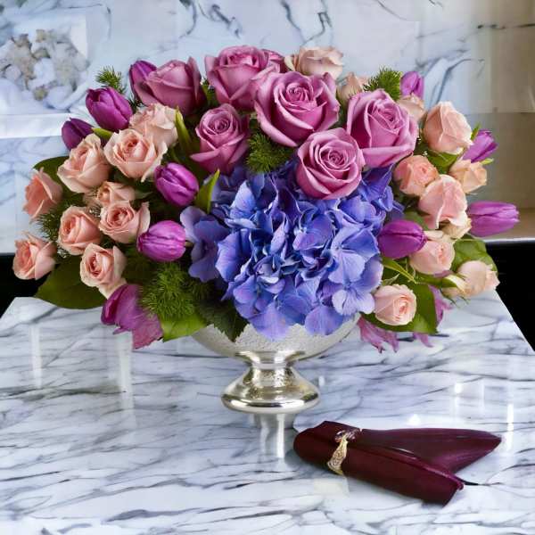 Pink roses and purple tulips arranged with blue hydrangea in a silver pedestal vase.
