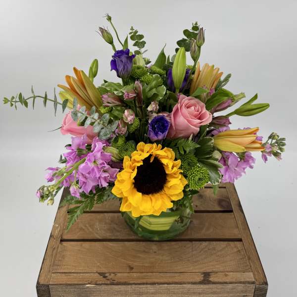Mixed arrangement of sunflowers, pink roses, lilies, and purple blooms in a round glass vase