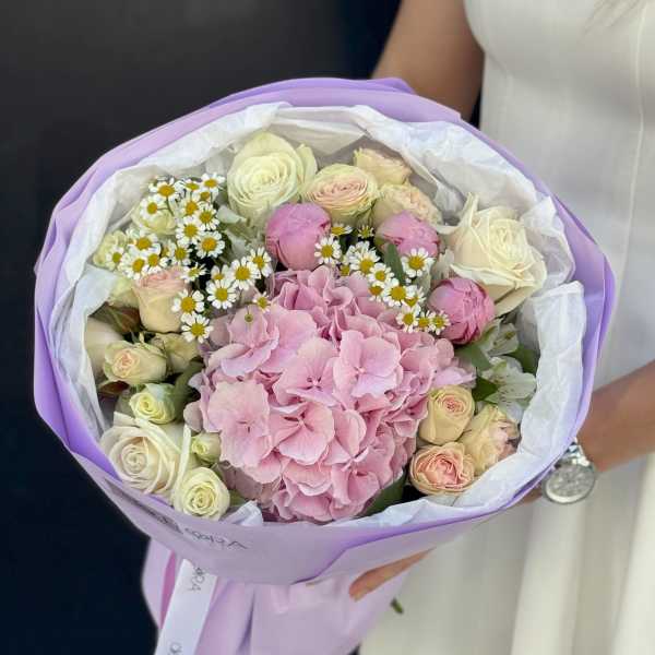 Bouquet of pink hydrangea, cream roses, and small white daisies wrapped in lavender paper