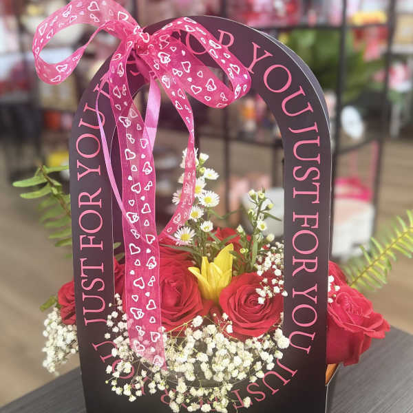 Pink roses and a yellow lily in a black gift bag with a heart ribbon.