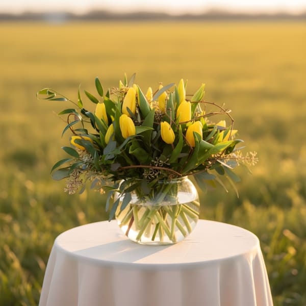 Yellow tulips with lush greenery arranged in a round glass vase on a draped table outdoors