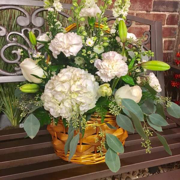 White floral arrangement in a wicker basket with lilies, hydrangea, and tall blooms