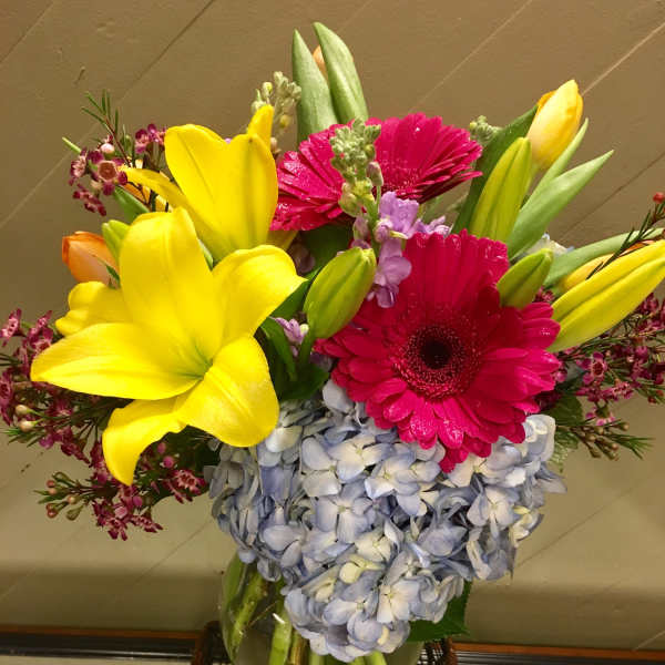 Bright mixed bouquet with yellow lilies, pink gerberas, and blue hydrangeas in a glass vase