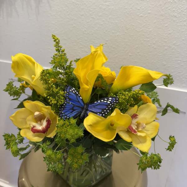 Yellow calla lilies and orchids in a glass vase with a blue butterfly accent