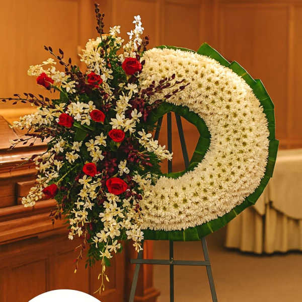 Large white crescent funeral wreath with red roses and white orchids on a metal easel stand.