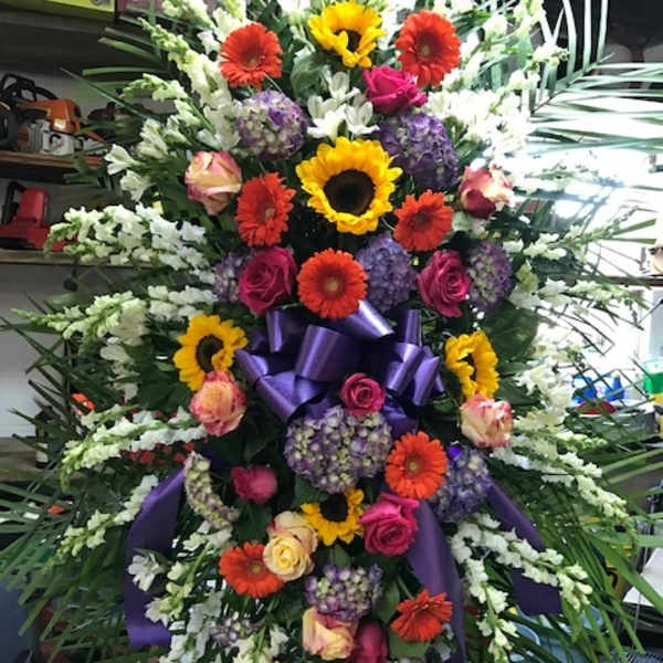 Large standing floral spray with sunflowers, gerbera daisies, roses, and purple ribbon