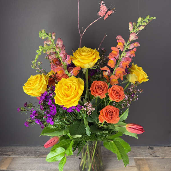 Tall mixed bouquet of yellow and orange roses with snapdragons, purple blooms and tulips in a clear glass vase